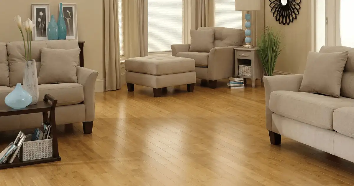 A living room with professionally installed bamboo flooring, beige sofas, a matching ottoman, a side table with a blue lamp, vases, framed pictures, and large windows with blinds and curtains in Las Vegas, NV